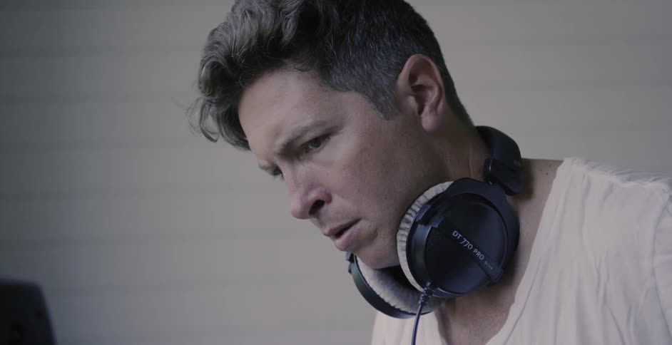 A man is wearing headphones around his neck as he stands indoors in daylight