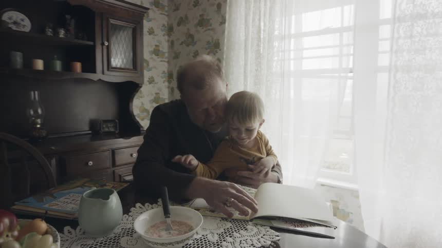 Grandfather is reading book to grandson at table in dining room
