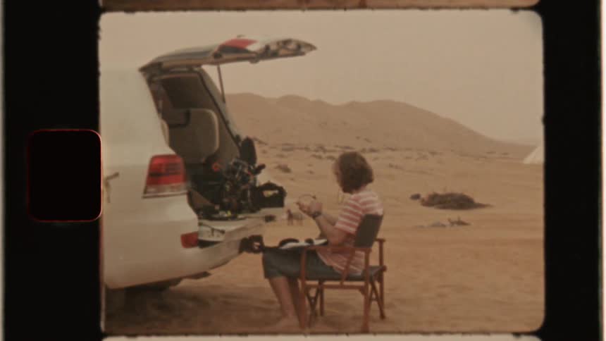 Woman in chair at back of truck in desert with movie camera