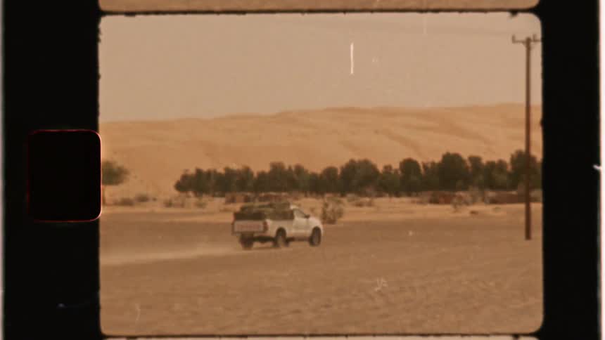 A pickup truck is speeding across the desert toward a building in daylight