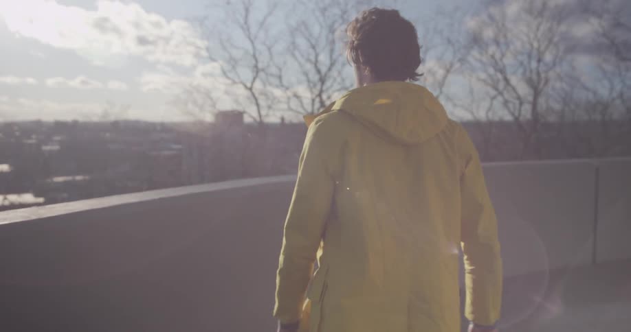 A man in a yellow jacket running his hand along a wall as he walks around it and then walks to a wall to look out over the city
