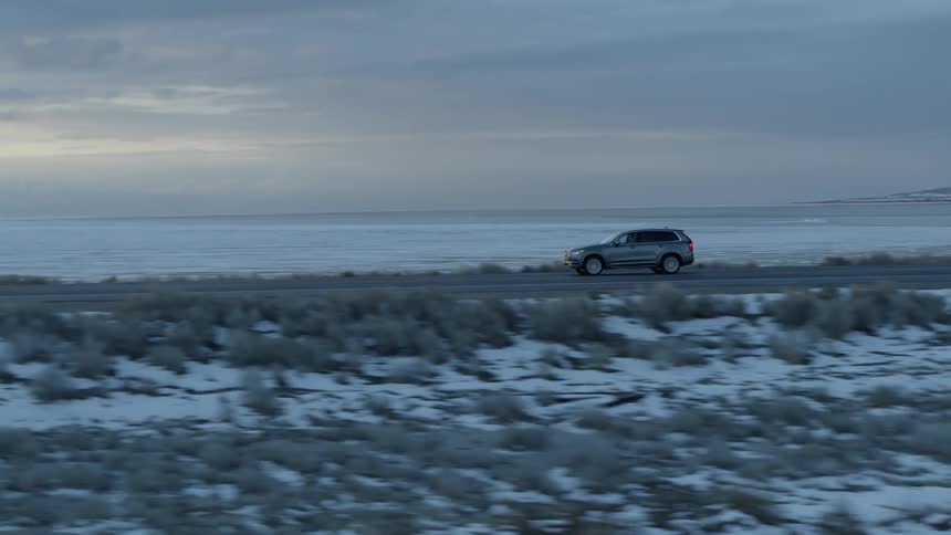 Vehicle drives on a road near snowy terrain and a body of water