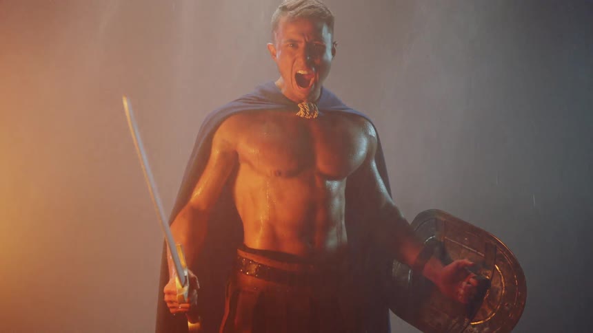 A man is yelling and brandishing a sword as he stands near a fire at dusk