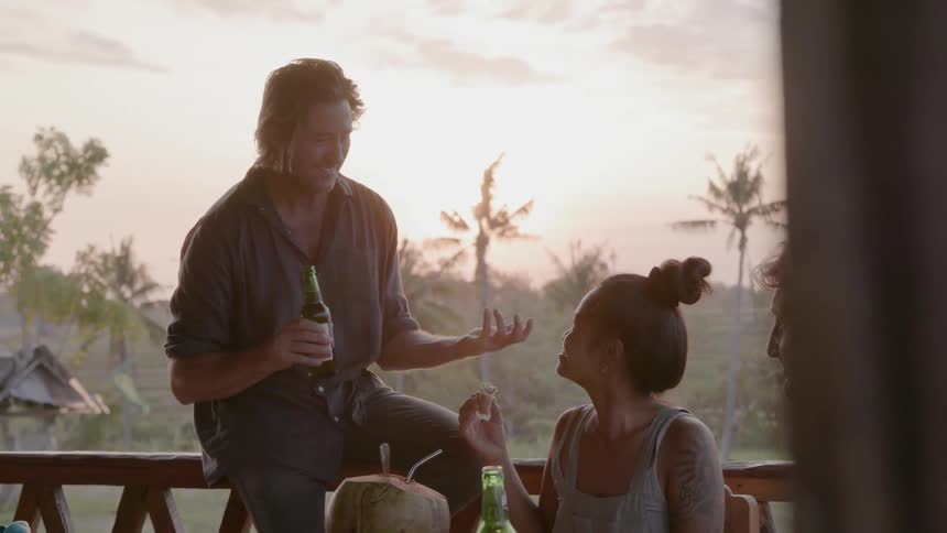 Man sits on a balcony railing talking to and drinking with a woman