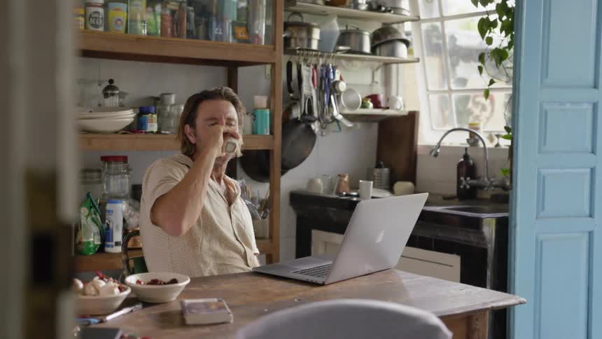 Man sits at a table in a kitchen looking at a laptop and drinking a beverage
