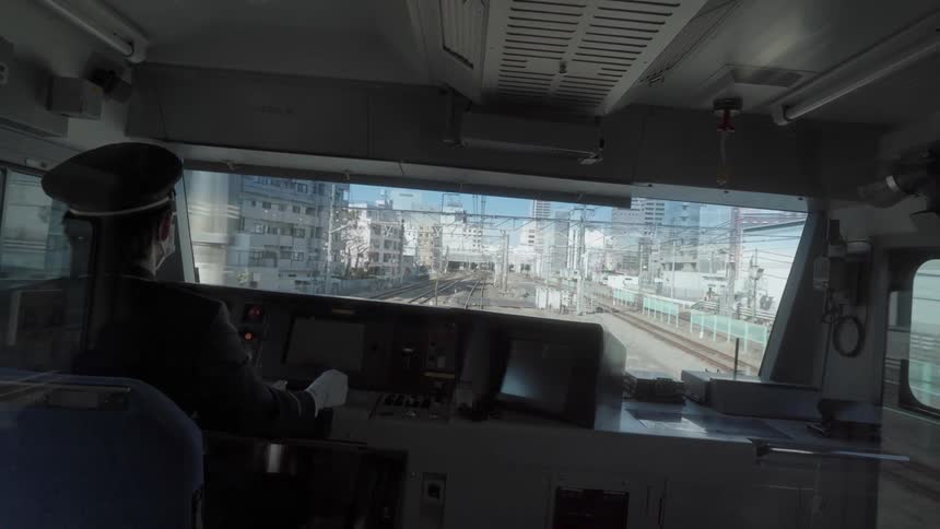 A man is driving a train through a city in daylight