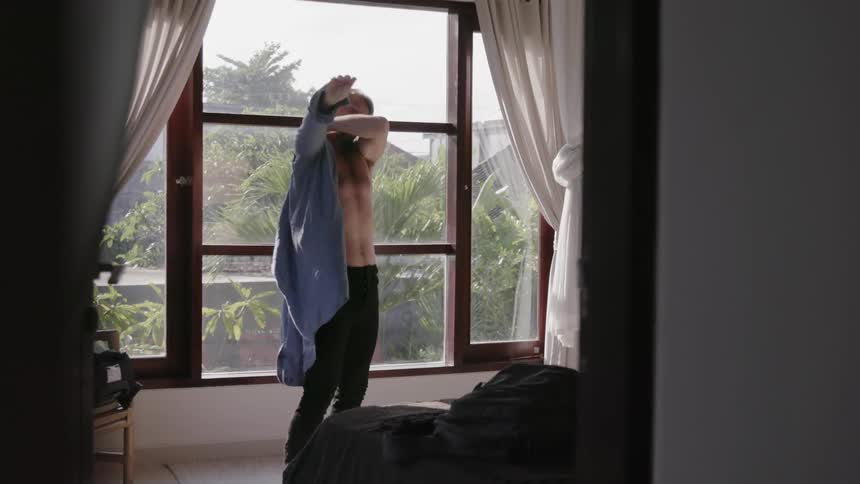 A man is putting on a shirt as he stands inside a room in daylight