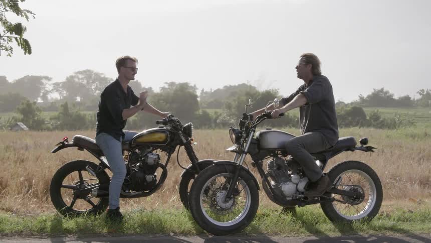 Two men sit on motorcycles in a field talking
