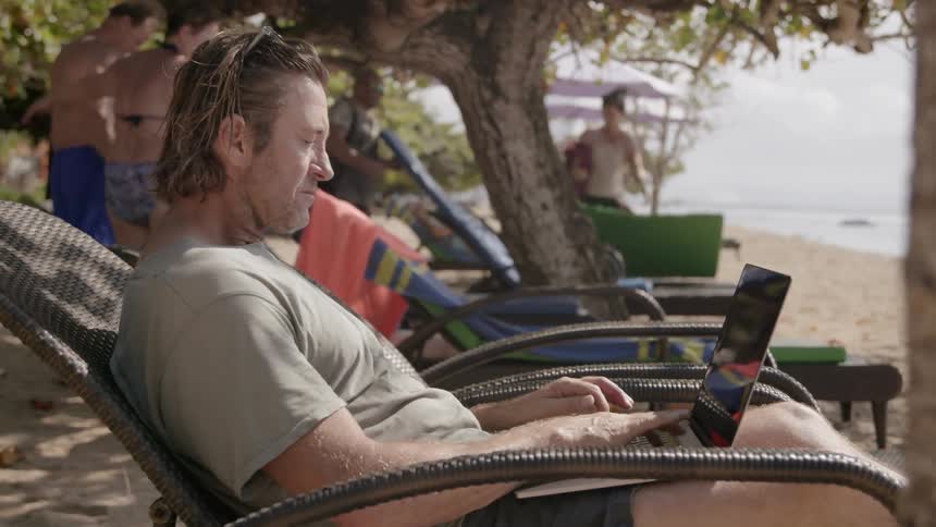 Man types on a laptop in a lawn chair on the beach