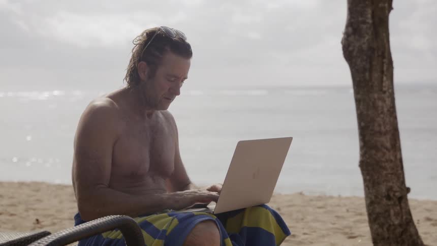Man sits on the beach typing on a laptop