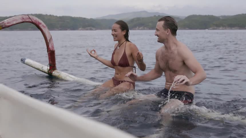 A man and woman are balancing on the edge of a boat in the ocean in daylight