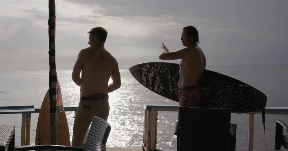A man holds a surfboard and talks to another on a deck on the ocean