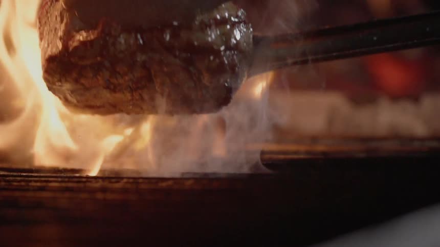 Tongs are flipping over a steak being grilled over flames outside at night