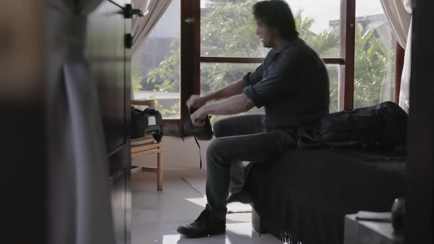A man sits on his bed and puts his boots on