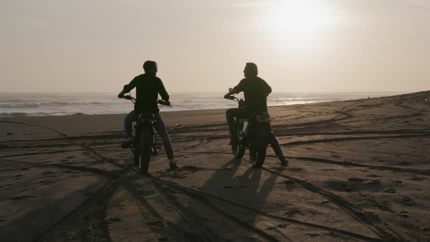 Two motorcyclists ride on a beach at sunset