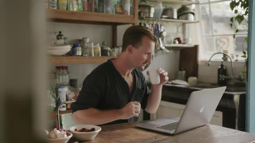 A man sits at a table drinking from a mug and working on a laptop