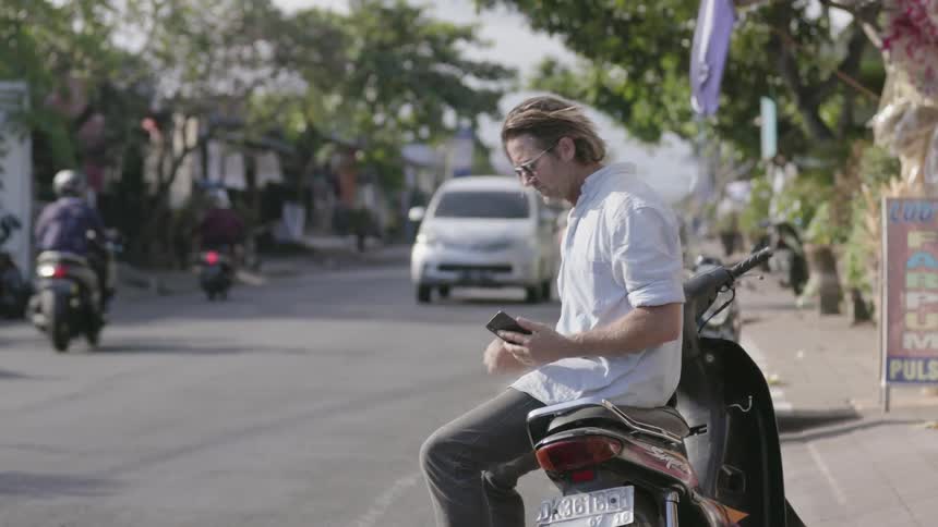 Man sits on a parked motorcycle on a city street looking at a phone