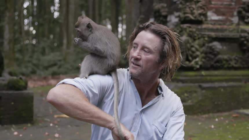 A man is holding a monkey on his shoulder as he sits outside in daylight