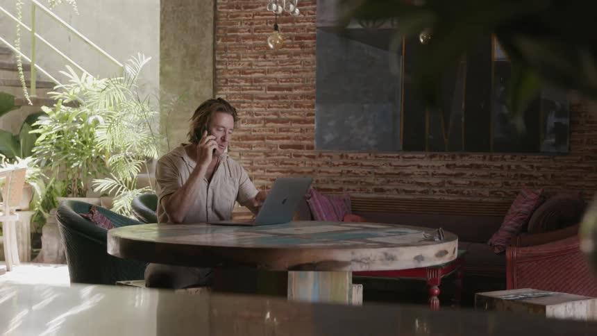 A man sitting at a table talks on the phone while looking at a laptop