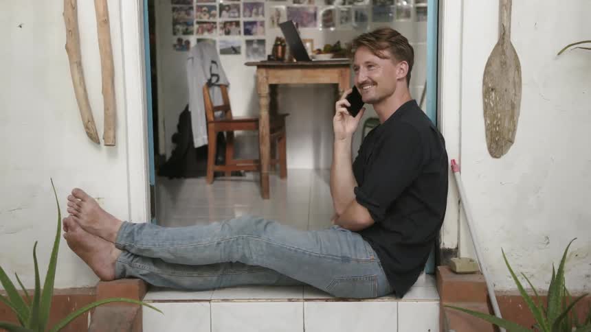 Man sits on a small porch talking on a phone and smiling