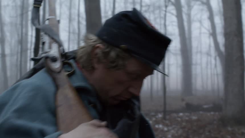 Man carrying a rifle puts a hat on his head as he walks in the forest