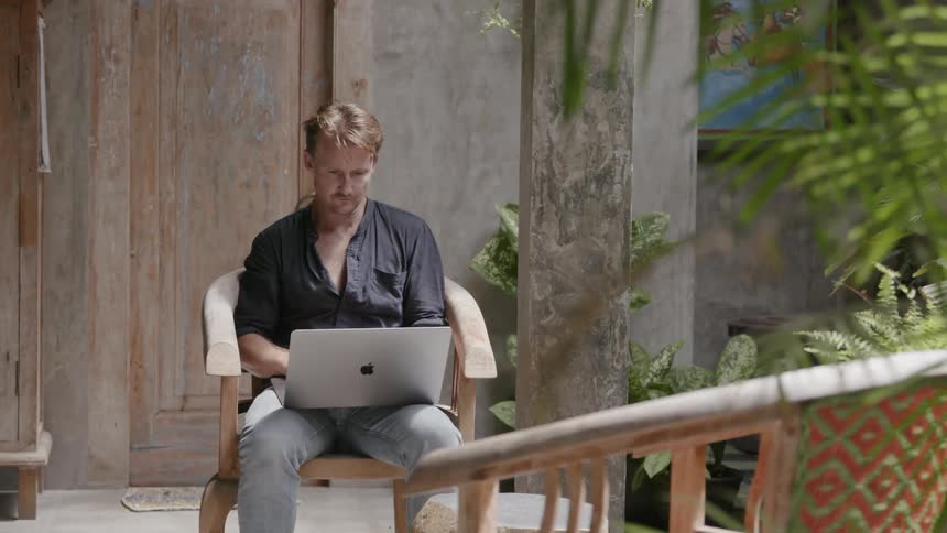 A man sits on a chair on a patio and works on a laptop