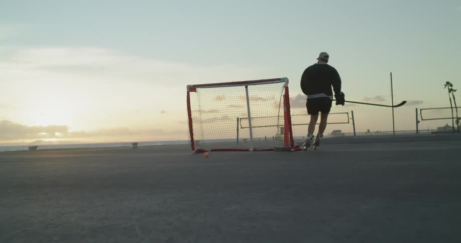 A man wearing roller blades shoots a ball into a net with a hockey stick
