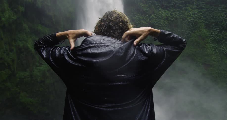 Person puts a hood on while standing near a waterfall