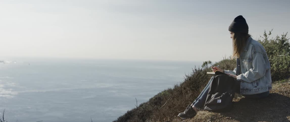 A woman sits on the hill by the ocean writing
