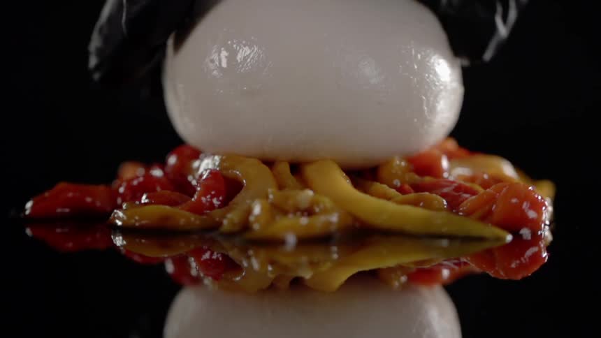 A gloved hand places a white bun on top of roasted peppers