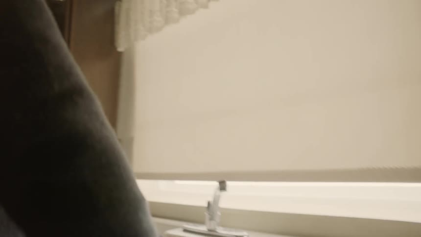Man leans over a kitchen sink looking at a window covered with a blind
