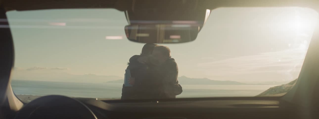 Two men hug in front of a parked car at sunset