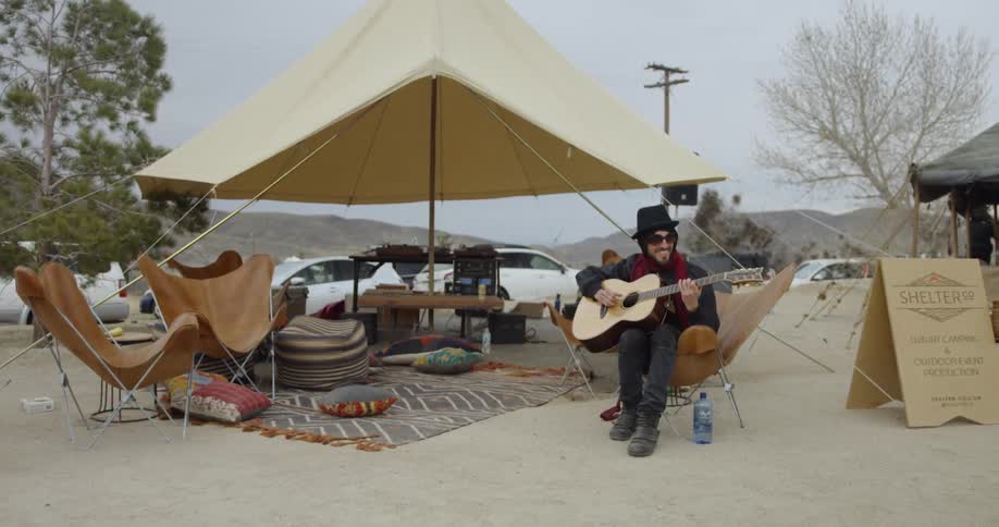 A man sits in a chair by the tent playing guitar