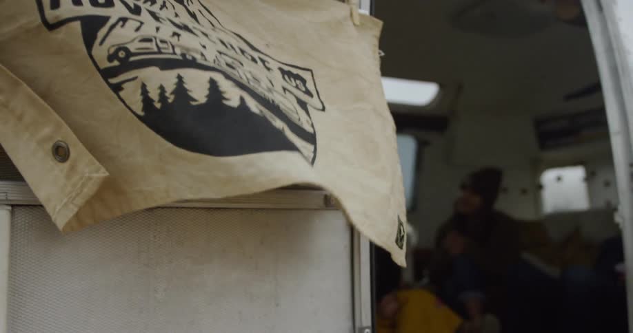 Flag waves in the wind on a camper door and people sit inside