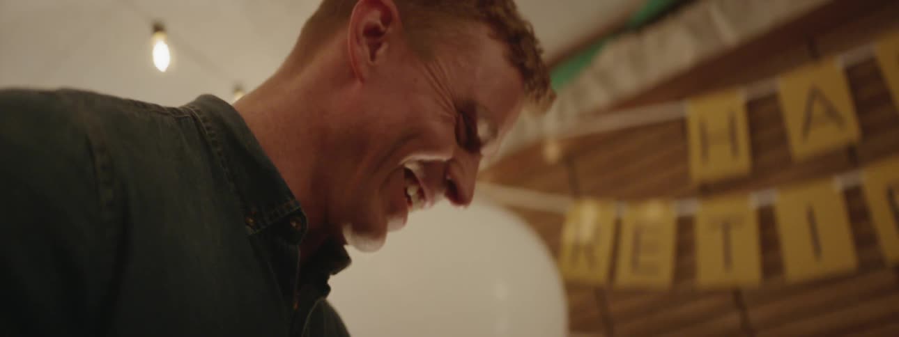 A man smiles and looks down as another cuts a cake at a party