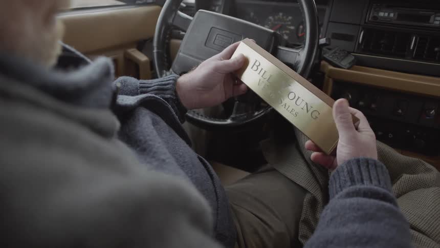 Man sits in a vehicle as he holds a rectangular block with his name on it