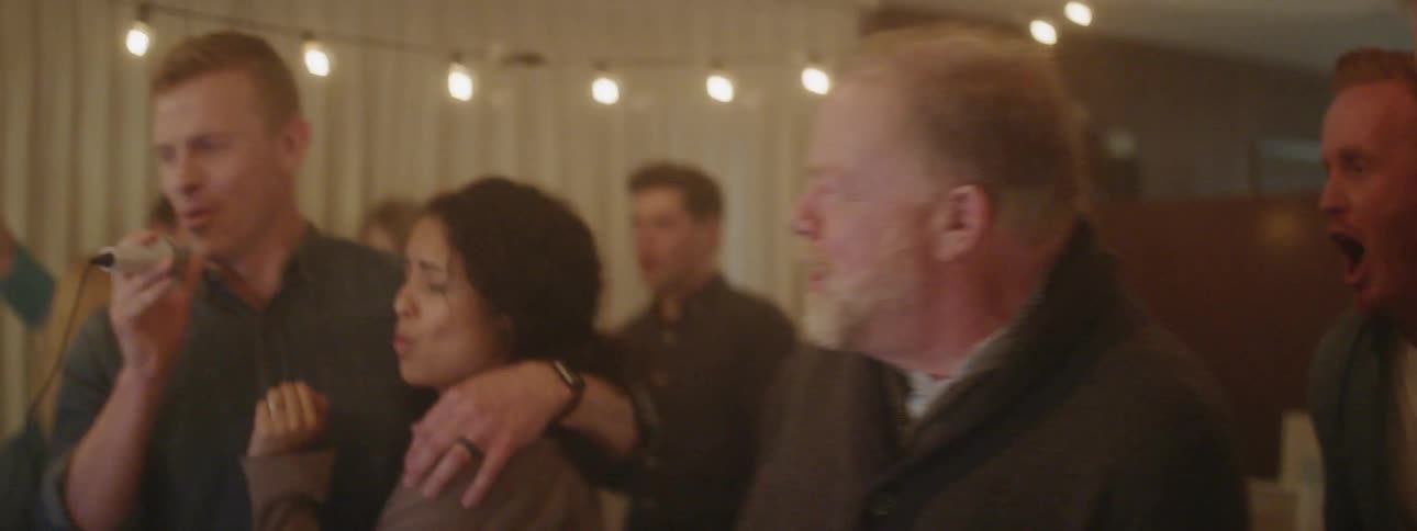 Man sings into a microphone as he has his arm around a woman