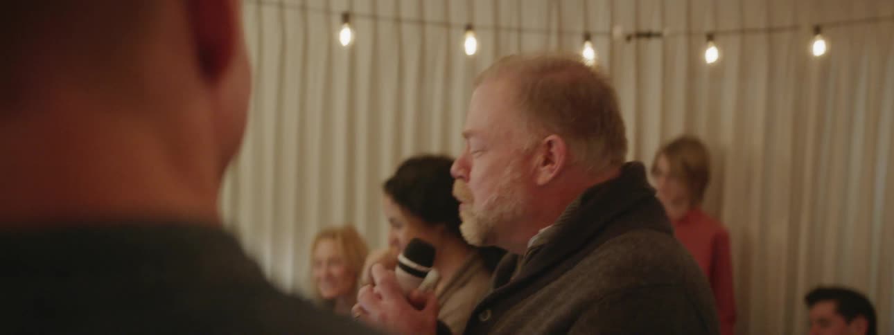 Man sings into a microphone as people are gathered around him