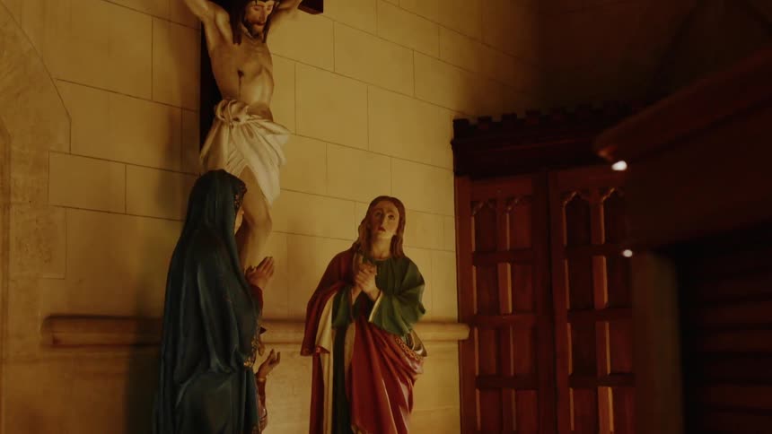 A crucifix hangs on the wall of a church with statues of mourners below