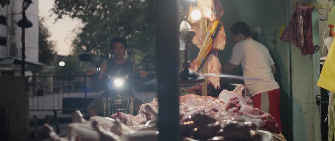 Low hanging fan spins around above a table of meat and a man stands behind the counter organizing
