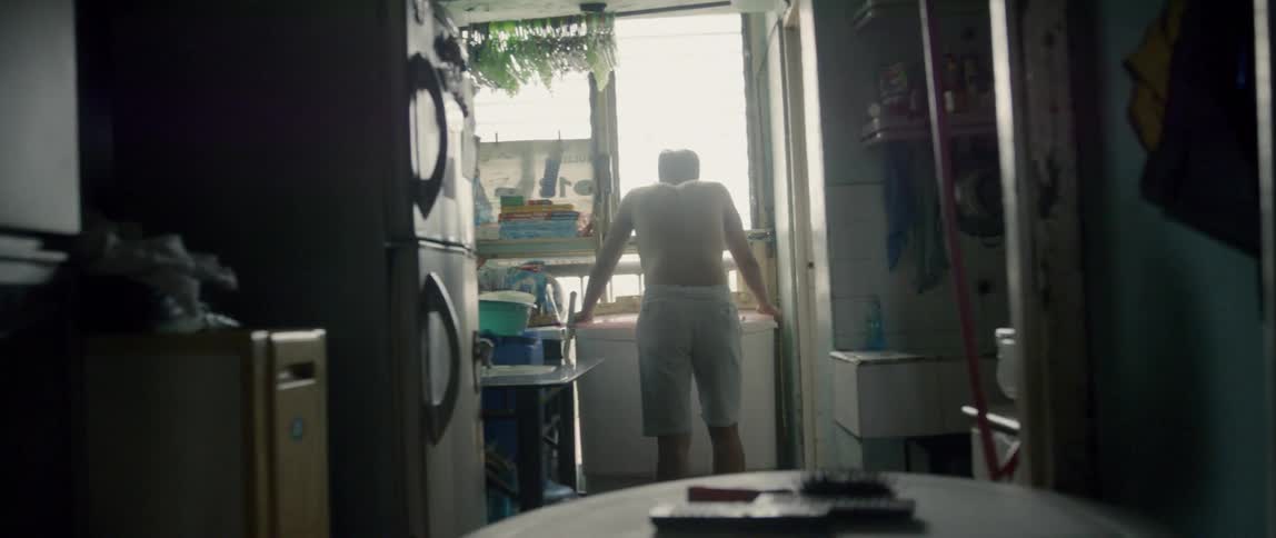 Shirtless man leans towards a window in the kitchen