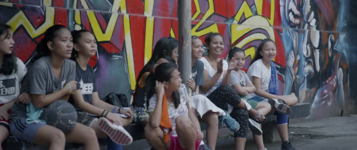 Teenagers and children sit in front of a graffiti covered wall clapping their hands