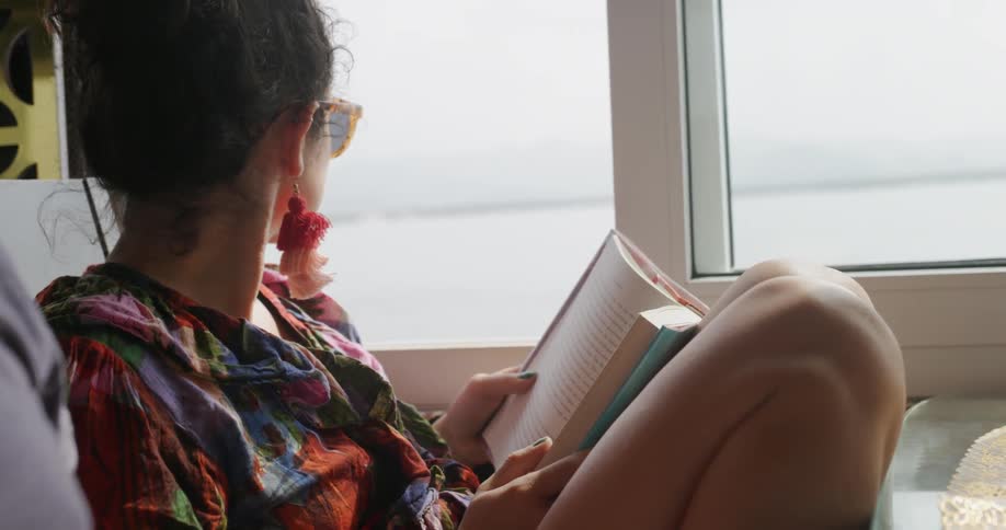 A woman sits next to a window by a body of water reading a book and then looking out