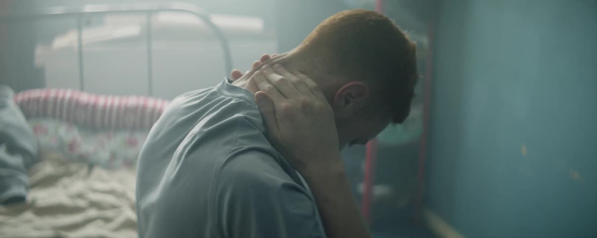 Man rubs the back of his neck as he sits on the edge of a bed