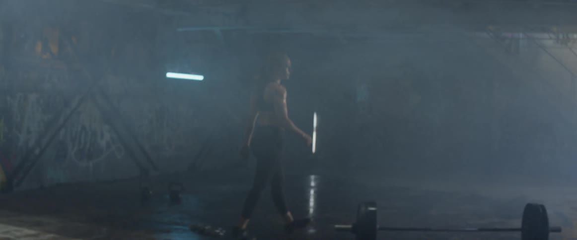 Woman walks in a dim room with a wet floor towards a barbell and picks it up