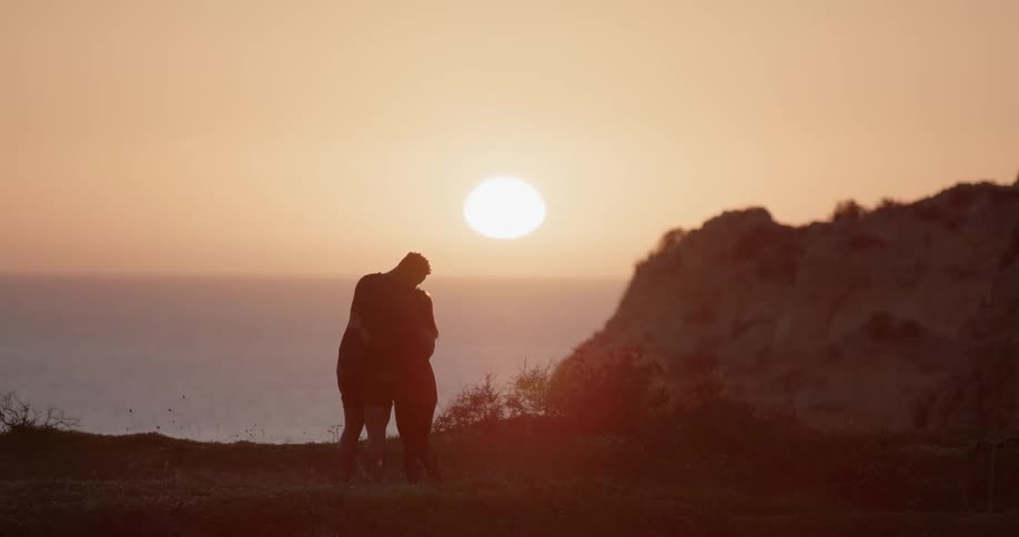 A man and woman stand on a beach at sunset and hug