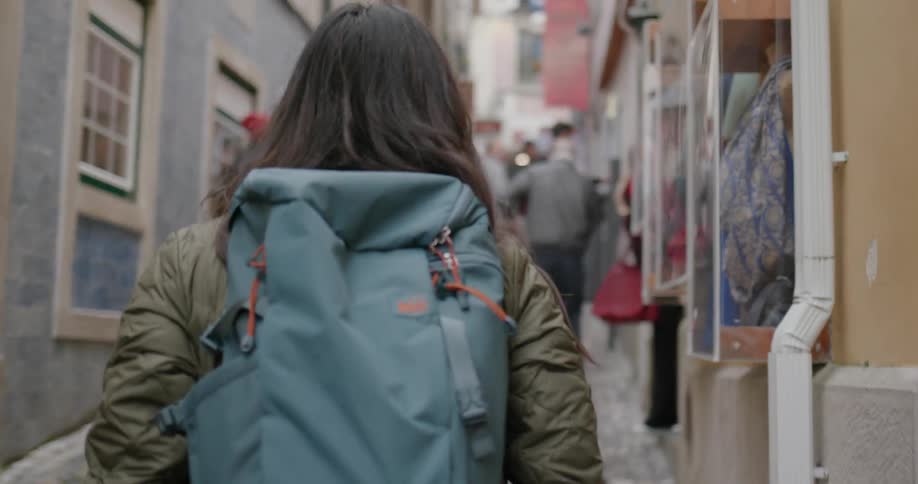 A woman wearing a backpack smiles and turns to walk down a busy alley at day time