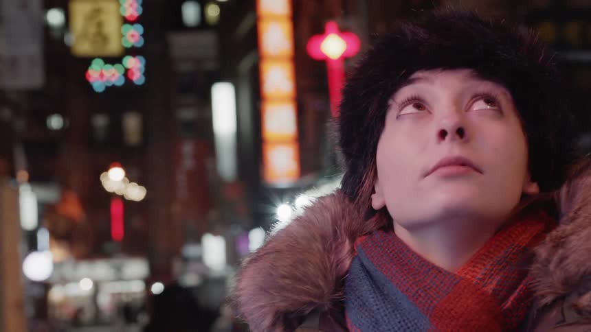 A woman is looking up at the city lights as she stands outside on a cold night