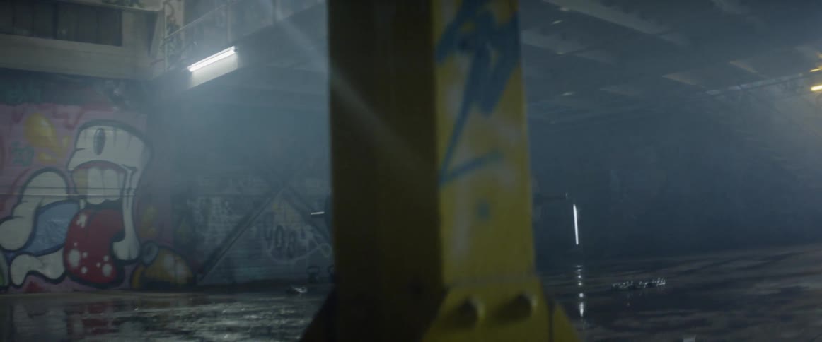 A man in a dark gym with graffiti on the walls lifts a large dumbbell