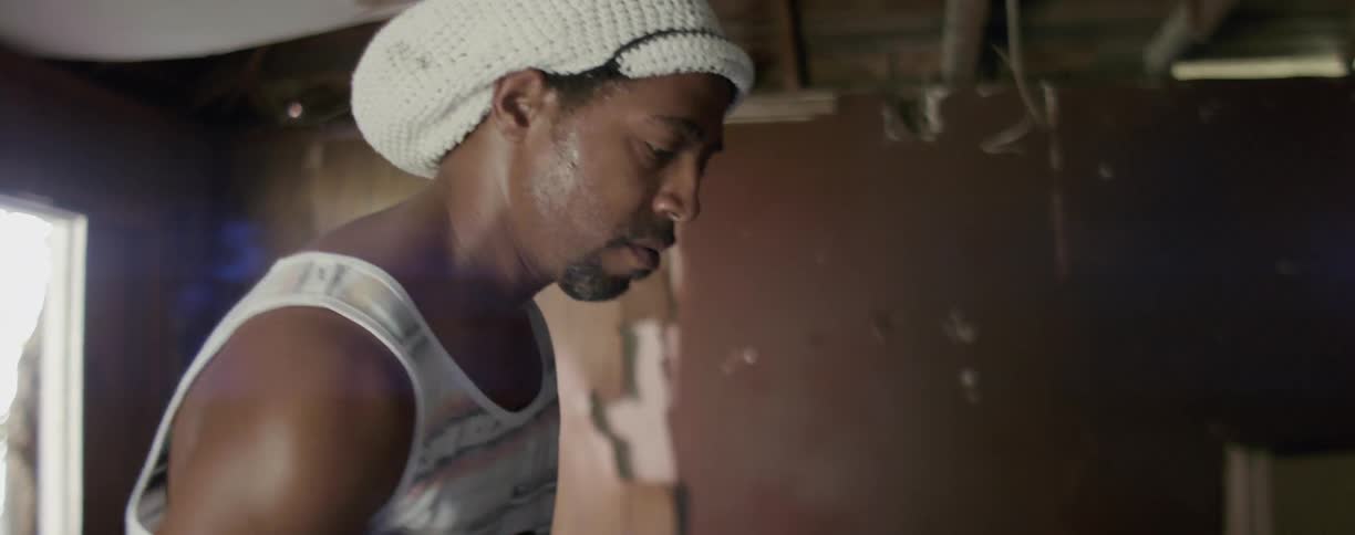A man is tapping his drums with a drumstick while standing inside in daylight